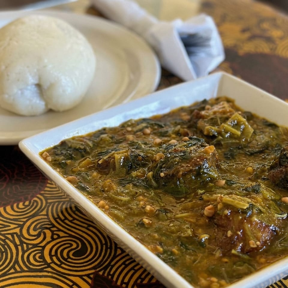A square dish of savory meatballs in rich sauce sits beside a round bread roll on a patterned restaurant table.